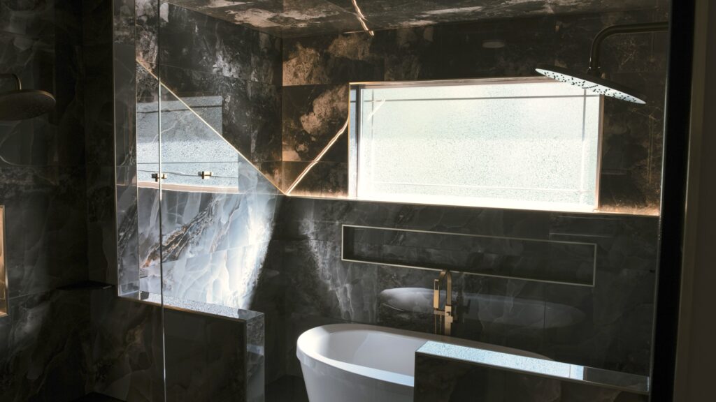 Alpenglow Builders LLC - A high-end modern bathroom with black marble walls, a large window, and a sleek white bathtub, showcasing luxury interior des.