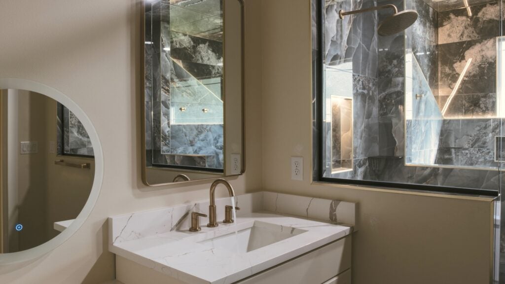 Alpenglow Builders LLC - A high-end bathroom vanity with a white marble countertop, modern gold fixtures, and large windows showcasing a natural outdo.