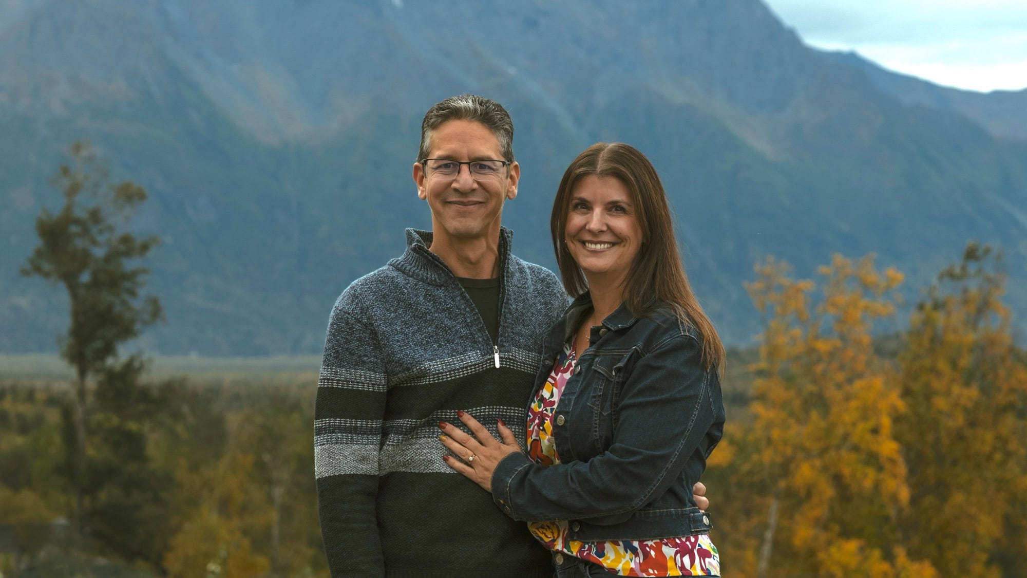 Alpenglow Builders LLC - Elegant couple standing outdoors with mountains and autumn trees in the background, showcasing luxury home architecture in Pa.