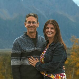 Alpenglow Builders LLC - A smiling couple standing outdoors with mountains and autumn trees in the background.